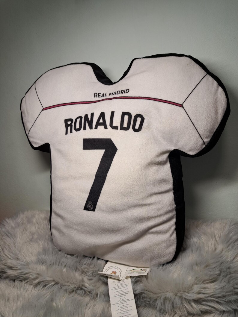 Puede incluir: Un coj&iacute;n con forma de camiseta blanca con bordes negros que presenta el texto REAL MADRID, el nombre RONALDO y el n&uacute;mero 7 en letras negras. El dise&ntilde;o incluye una fina l&iacute;nea horizontal roja en el pecho y un peque&ntilde;o logotipo en la base.