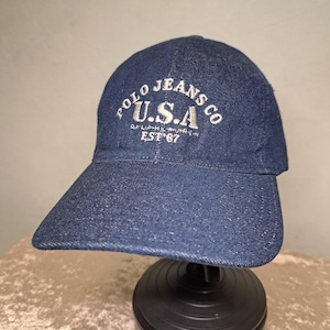 Puede incluir: Gorra de béisbol de mezclilla azul oscuro con visera curva. La gorra presenta texto bordado en blanco que dice "POLO JEANS CO U.S.A." y "EST '67". El sombrero se exhibe en un soporte negro.