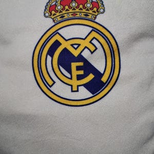 Puede incluir: Una superficie de tela blanca con el logotipo del club de f&uacute;tbol Real Madrid. El emblema consiste en un borde circular dorado con las letras MCF entrelazadas en oro, una franja diagonal azul oscuro y una corona dorada con detalles rojos y azules.