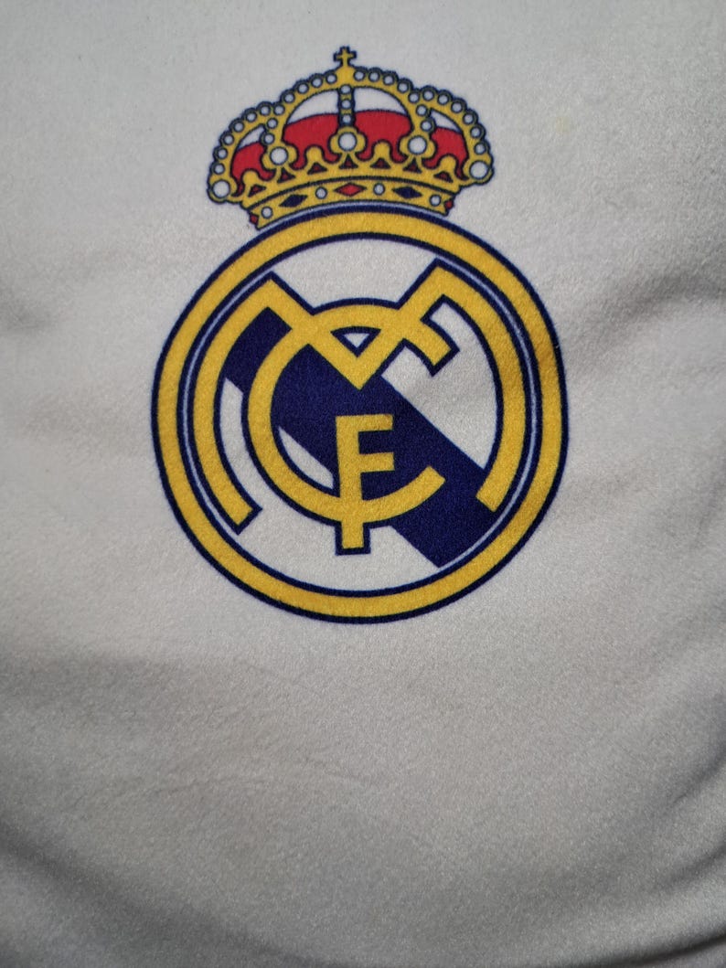 Puede incluir: Una superficie de tela blanca con el logotipo del club de f&uacute;tbol Real Madrid. El emblema consiste en un borde circular dorado con las letras MCF entrelazadas en oro, una franja diagonal azul oscuro y una corona dorada con detalles rojos y azules.