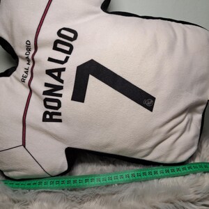 Puede incluir: Un coj&iacute;n con forma de camiseta blanca que presenta el texto REAL MADRID, RONALDO y el n&uacute;mero 7 en negro. Una cinta m&eacute;trica verde est&aacute; colocada a lo largo de la parte inferior, mostrando una longitud de unos 44 cent&iacute;metros para la base.