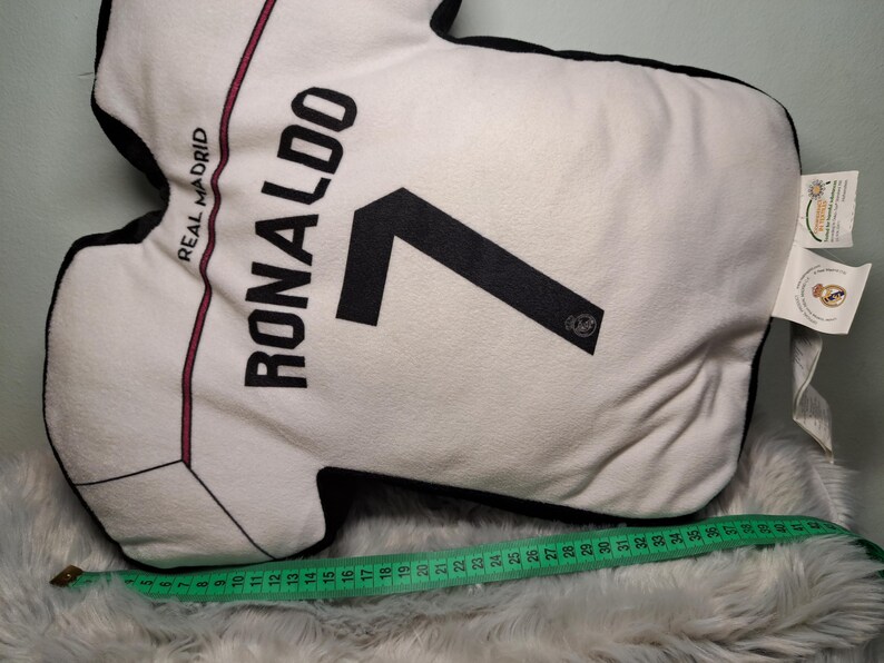Puede incluir: Un coj&iacute;n con forma de camiseta blanca que presenta el texto REAL MADRID, RONALDO y el n&uacute;mero 7 en negro. Una cinta m&eacute;trica verde est&aacute; colocada a lo largo de la parte inferior, mostrando una longitud de unos 44 cent&iacute;metros para la base.