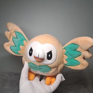 May include: A plush toy shaped like a round bird with tan fur, green wing accents, a white facial disc, and orange feet. The character has black eyes with a stern expression, a small beak, and a green bow-like feature on its chest, crafted from soft fabric.