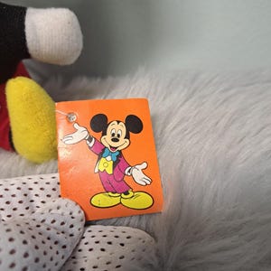 Pu&ograve; includere: Un peluche vintage di Topolino con un'etichetta con un'immagine a cartoni animati di Mickey con una giacca viola, scarpe gialle e un papillon blu. L'etichetta &egrave; arancione. Il peluche ha orecchie nere, viso bianco e guanti gialli.