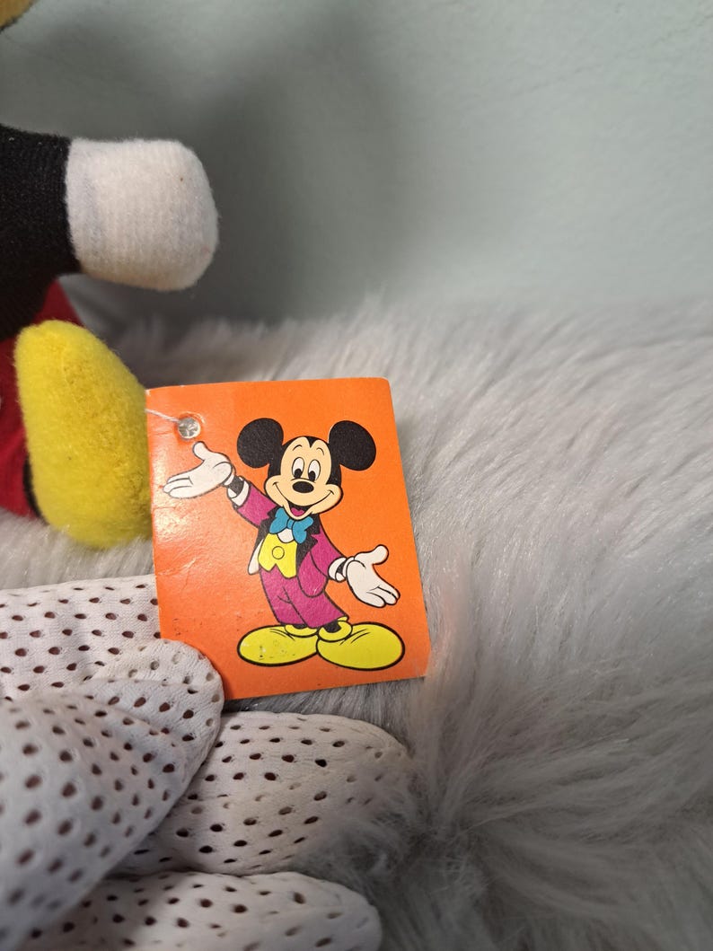 Pu&ograve; includere: Un peluche vintage di Topolino con un'etichetta con un'immagine a cartoni animati di Mickey con una giacca viola, scarpe gialle e un papillon blu. L'etichetta &egrave; arancione. Il peluche ha orecchie nere, viso bianco e guanti gialli.