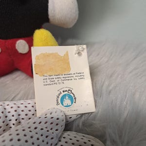 Pu&ograve; includere: Un peluche vintage di Topolino con un'etichetta. Il peluche ha un corpo nero, pantaloncini rossi, scarpe gialle e guanti bianchi. L'etichetta recita "Disneyland Walt Disney World" e include testo sulle normative di sicurezza.