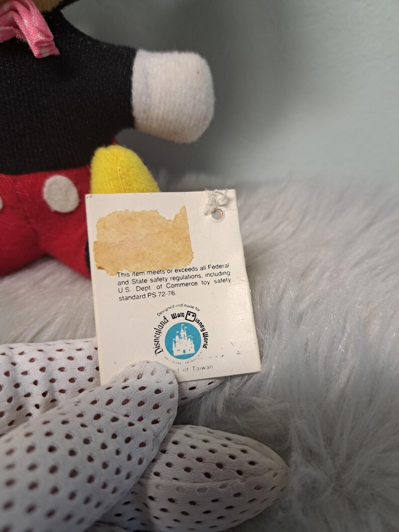 Pu&ograve; includere: Un peluche vintage di Topolino con un'etichetta. Il peluche ha un corpo nero, pantaloncini rossi, scarpe gialle e guanti bianchi. L'etichetta recita "Disneyland Walt Disney World" e include testo sulle normative di sicurezza.