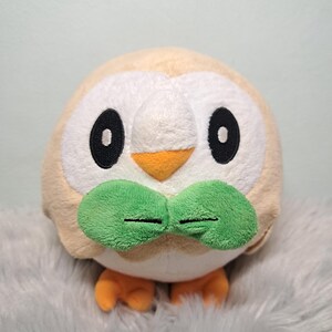 May include: A round, plush toy shaped like an owl with tan and white fur. It features large black oval eyes, a small orange triangular beak, and two prominent green leaf-shaped appendages below the beak. Two small orange feet are visible at the base.