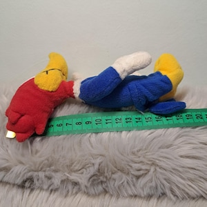 May include: A plush toy featuring a red head, yellow beak, blue body, and white hands, lying horizontally next to a green measuring tape. The toy measures approximately 15 centimeters in length from the beak to the feet as shown on the tape.