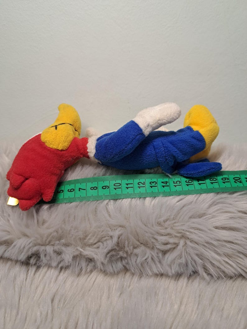 May include: A plush toy featuring a red head, yellow beak, blue body, and white hands, lying horizontally next to a green measuring tape. The toy measures approximately 15 centimeters in length from the beak to the feet as shown on the tape.