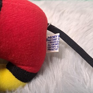 Pu&ograve; includere: Primo piano di un peluche di Topolino. Il giocattolo presenta un corpo rosso, dettagli gialli e una coda nera. Un'etichetta con testo &egrave; attaccata. Il peluche &egrave; appoggiato su una superficie bianca e soffice.