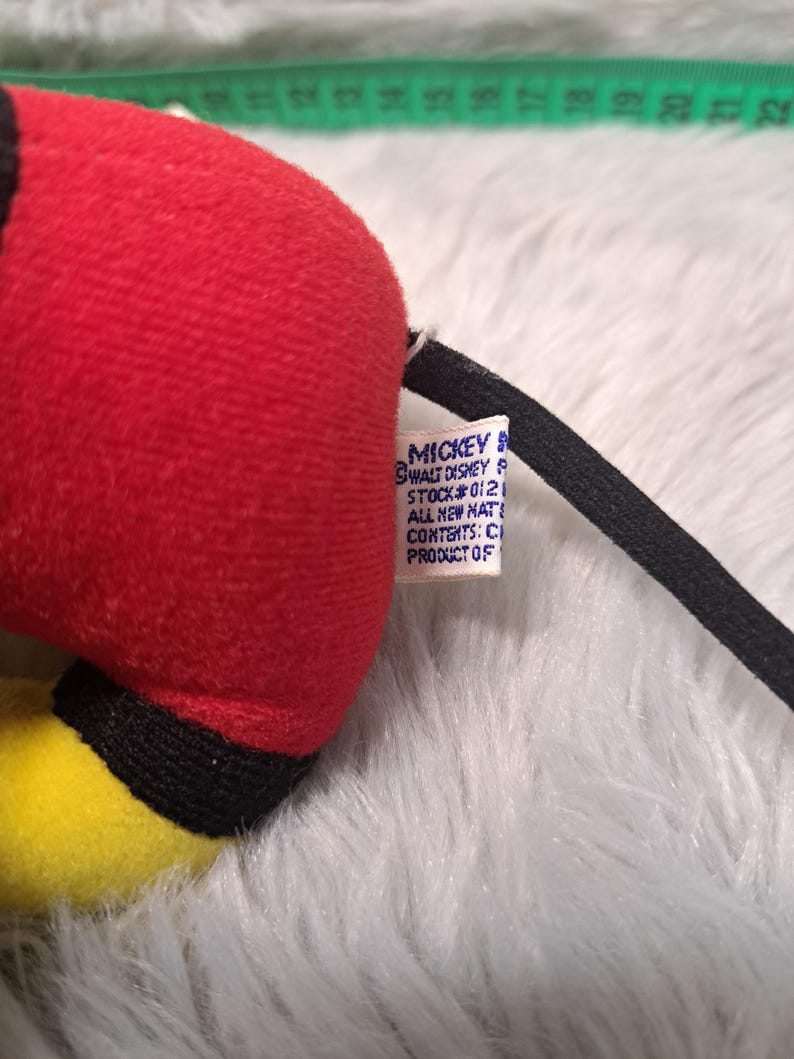 Pu&ograve; includere: Primo piano di un peluche di Topolino. Il giocattolo presenta un corpo rosso, dettagli gialli e una coda nera. Un'etichetta con testo &egrave; attaccata. Il peluche &egrave; appoggiato su una superficie bianca e soffice.