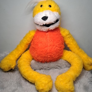 May include: A plush toy featuring a bright yellow body with long limbs, a vibrant orange torso, and a white muzzle. The character has black eyes, a black nose, a wide black mouth, and a tuft of white hair on top of its head, all made from soft, textured fabric.