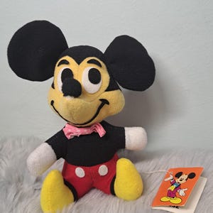 Pu&ograve; includere: Un peluche vintage di Topolino. Il peluche ha un corpo, orecchie e braccia nere, con pantaloncini rossi, piedi gialli e un papillon rosa. Un'etichetta con un'illustrazione di Topolino &egrave; attaccata.