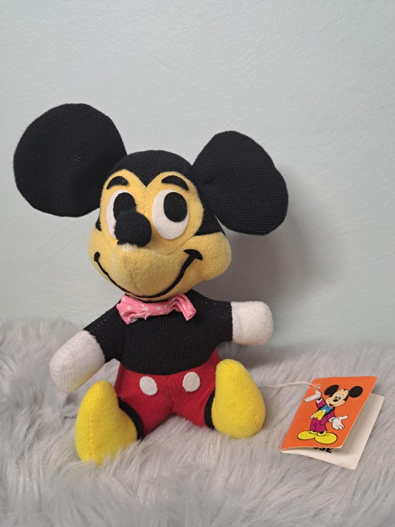 Pu&ograve; includere: Un peluche vintage di Topolino. Il peluche ha un corpo, orecchie e braccia nere, con pantaloncini rossi, piedi gialli e un papillon rosa. Un'etichetta con un'illustrazione di Topolino &egrave; attaccata.
