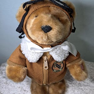 May include: A plush teddy bear wearing a brown aviator jacket with a white faux fur collar and a black aviator cap. The jacket has a zipper and a patch that reads "2003". The bear has a black nose and is standing upright.
