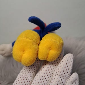 May include: A plush toy featuring two large, rounded yellow feet with stitched details, connected to a blue fabric section. The toy has a small blue tail extending upward from the center, with a glimpse of red fabric visible behind the blue base.