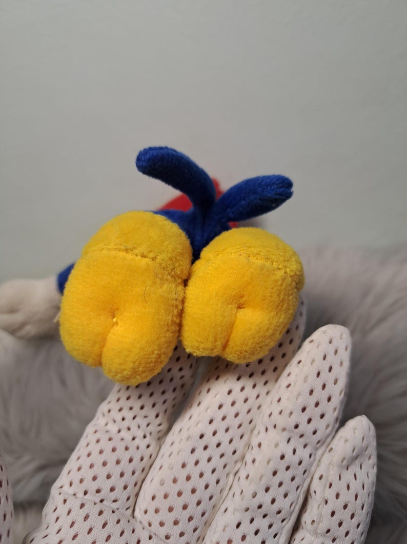 May include: A plush toy featuring two large, rounded yellow feet with stitched details, connected to a blue fabric section. The toy has a small blue tail extending upward from the center, with a glimpse of red fabric visible behind the blue base.
