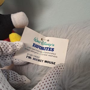 Pu&ograve; includere: Primo piano di un peluche di Topolino con un'etichetta che recita "Walt Disney's FAVORITES". L'etichetta dice anche "I'M: MICKEY MOUSE". Il giocattolo ha guanti bianchi con piccoli fori.