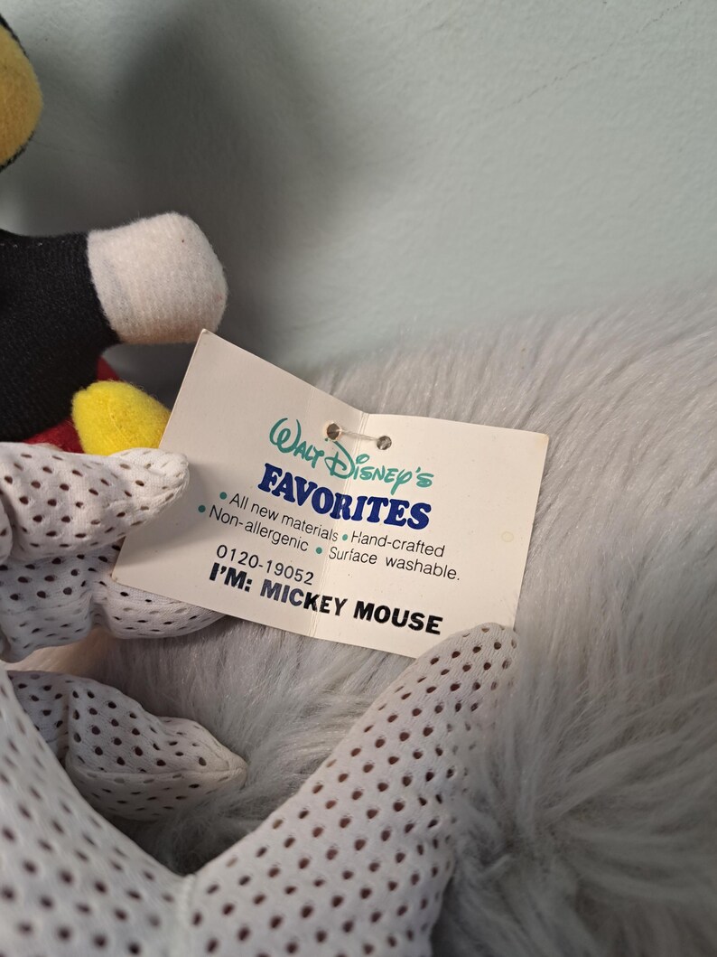 Pu&ograve; includere: Primo piano di un peluche di Topolino con un'etichetta che recita "Walt Disney's FAVORITES". L'etichetta dice anche "I'M: MICKEY MOUSE". Il giocattolo ha guanti bianchi con piccoli fori.