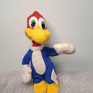 May include: A plush toy of a cartoon woodpecker with a red head, a yellow beak, and large white eyes with green irises. The character has a blue body with a white chest, white hands, and yellow feet. A small white fabric tag is attached to the side.