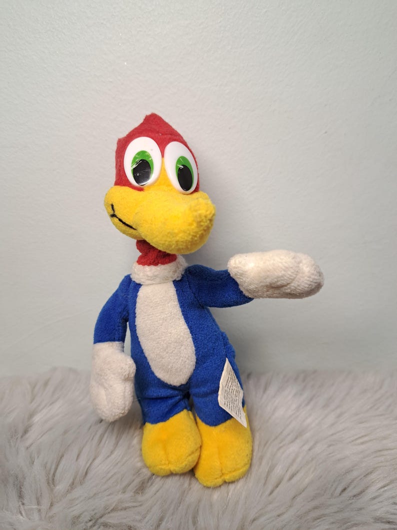 May include: A plush toy of a cartoon woodpecker with a red head, a yellow beak, and large white eyes with green irises. The character has a blue body with a white chest, white hands, and yellow feet. A small white fabric tag is attached to the side.