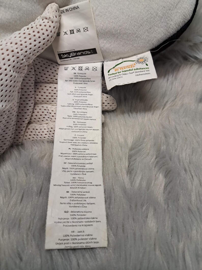 Puede incluir: Una etiqueta de cuidado de tela blanca unida a un art&iacute;culo de felpa, con el logotipo de SkyBrands y el texto MADE IN CHINA. Incluye m&uacute;ltiples s&iacute;mbolos de lavado y una etiqueta secundaria con la certificaci&oacute;n Oeko-Tex Standard 100.