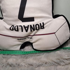 Puede incluir: Un coj&iacute;n blanco y negro con forma de camiseta de f&uacute;tbol con el n&uacute;mero 7, el nombre RONALDO y el texto REAL MADRID impresos. Una cinta m&eacute;trica verde est&aacute; colocada en la parte inferior, mostrando una anchura de 63,5 cm.