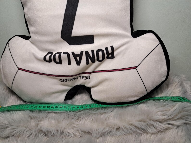 Puede incluir: Un coj&iacute;n blanco y negro con forma de camiseta de f&uacute;tbol con el n&uacute;mero 7, el nombre RONALDO y el texto REAL MADRID impresos. Una cinta m&eacute;trica verde est&aacute; colocada en la parte inferior, mostrando una anchura de 63,5 cm.