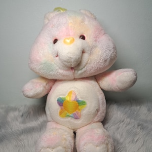Peut inclure: Une peluche en forme d'ours avec un motif tie-dye pastel doux dans des tons de rose, jaune et bleu. L'ours possède un nez en forme de cœur jaune et un écusson central sur le ventre représentant une étoile multicolore avec un cœur jaune.