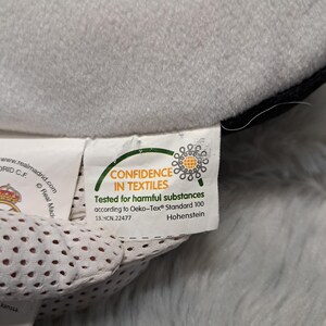 Puede incluir: Una etiqueta de tela blanca unida a un objeto de felpa gris. La etiqueta muestra el logotipo Oeko-Tex Confidence in Textiles, el texto Tested for harmful substances according to Oeko-Tex Standard 100 y el c&oacute;digo 13.HCN.22477 con Hohenstein.