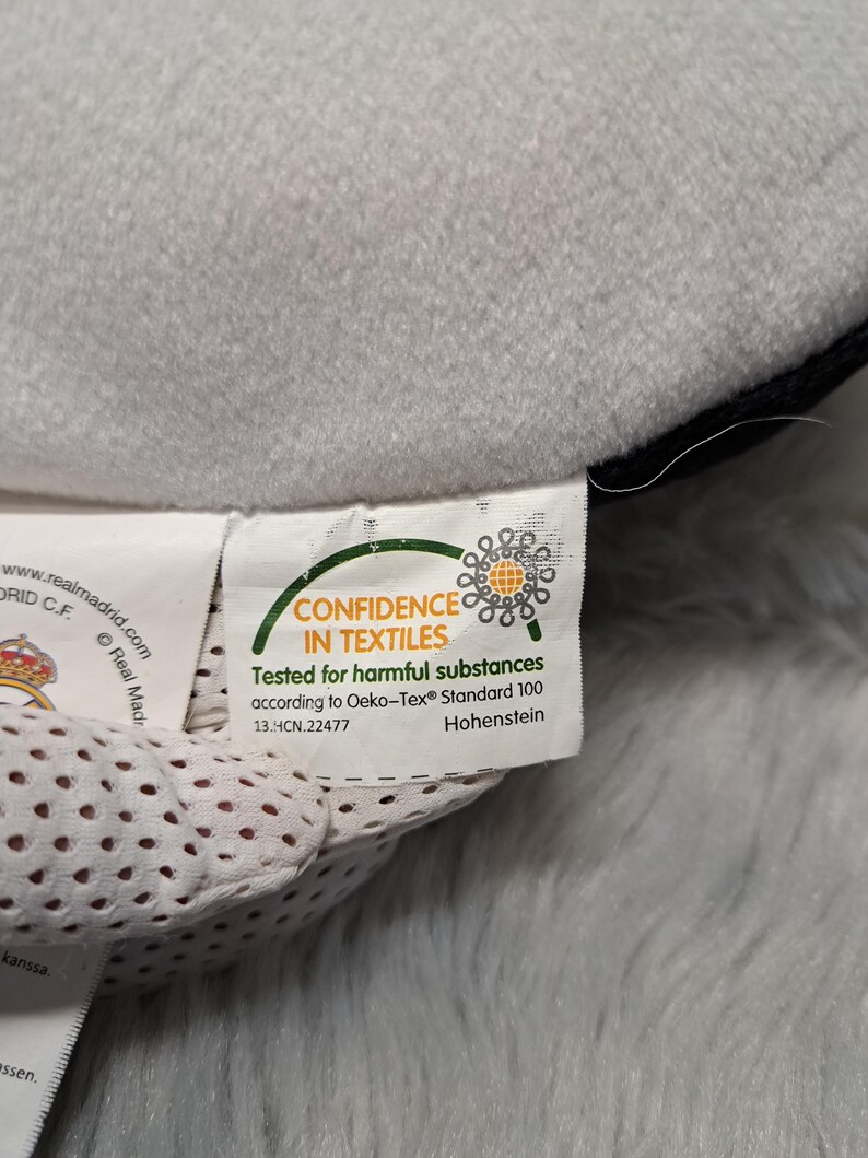 Puede incluir: Una etiqueta de tela blanca unida a un objeto de felpa gris. La etiqueta muestra el logotipo Oeko-Tex Confidence in Textiles, el texto Tested for harmful substances according to Oeko-Tex Standard 100 y el c&oacute;digo 13.HCN.22477 con Hohenstein.