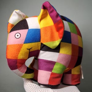Puede incluir: Un juguete de elefante de peluche colorido con un diseño de patchwork. El elefante presenta una variedad de colores, incluyendo naranja, amarillo, rosa, azul y morado. El juguete tiene una cara sonriente y orejas grandes.