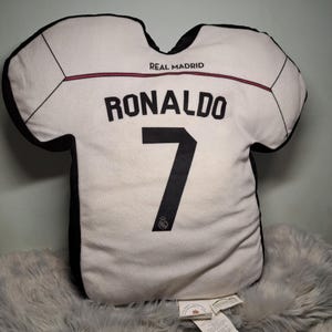 Puede incluir: Un coj&iacute;n decorativo con forma de camiseta blanca con bordes negros y el texto REAL MADRID. La parte posterior muestra el nombre RONALDO en letras negras sobre el n&uacute;mero 7, con un peque&ntilde;o logotipo negro debajo del n&uacute;mero en la tela.