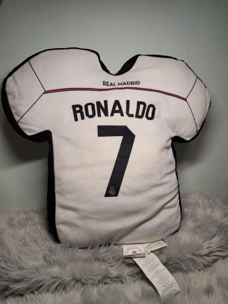 Puede incluir: Un coj&iacute;n decorativo con forma de camiseta blanca con bordes negros y el texto REAL MADRID. La parte posterior muestra el nombre RONALDO en letras negras sobre el n&uacute;mero 7, con un peque&ntilde;o logotipo negro debajo del n&uacute;mero en la tela.