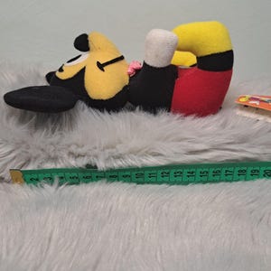 Pu&ograve; includere: Un peluche di Topolino sdraiato su un fianco. Il giocattolo ha una testa gialla, orecchie nere e un corpo nero. I pantaloncini sono rossi e i piedi sono neri. Un metro a nastro verde &egrave; accanto al giocattolo.