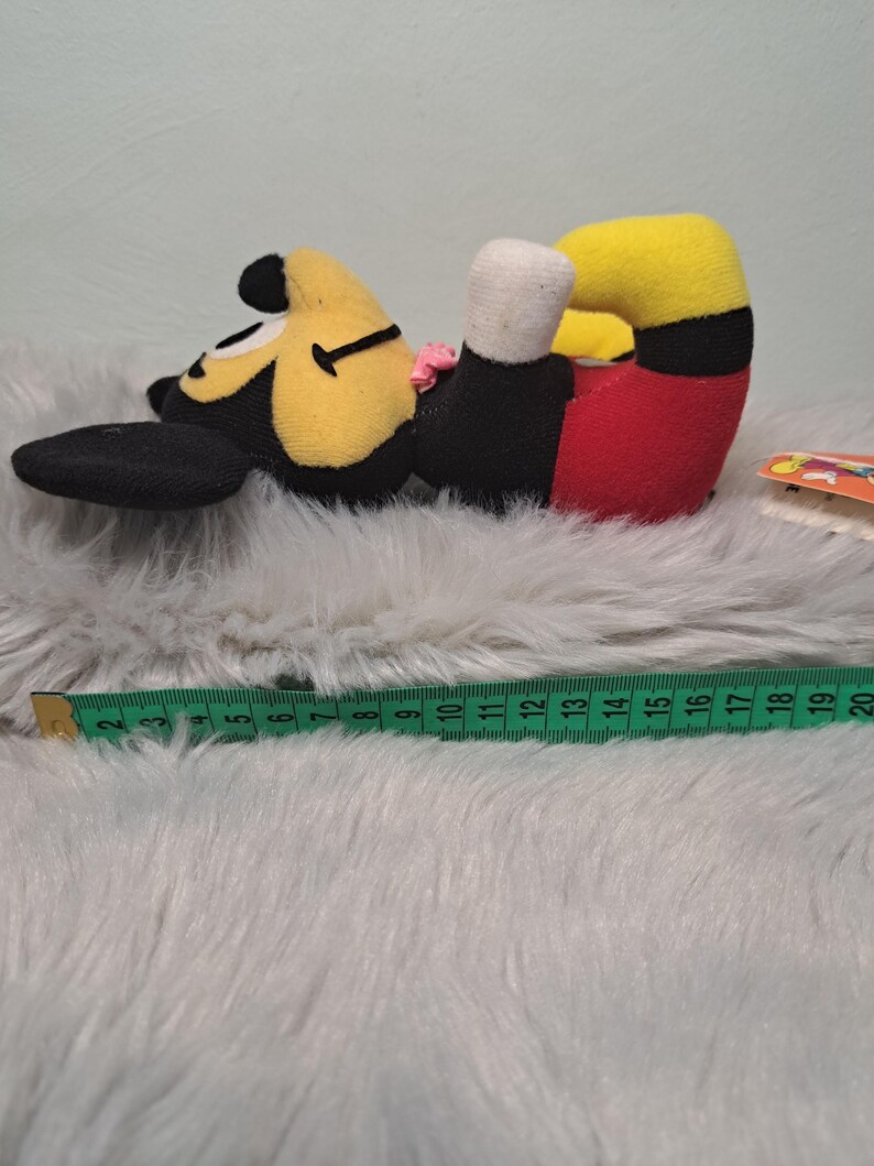 Pu&ograve; includere: Un peluche di Topolino sdraiato su un fianco. Il giocattolo ha una testa gialla, orecchie nere e un corpo nero. I pantaloncini sono rossi e i piedi sono neri. Un metro a nastro verde &egrave; accanto al giocattolo.