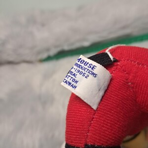 Pu&ograve; includere: Primo piano del braccio rosso di un peluche con polsino nero e finiture gialle. Un'etichetta bianca &egrave; attaccata, che mostra il testo tra cui "HOUSE PRODUCTIONS" e "MATERIAL COTTON TAIWAN". Lo sfondo &egrave; un grigio morbido e sfocato.