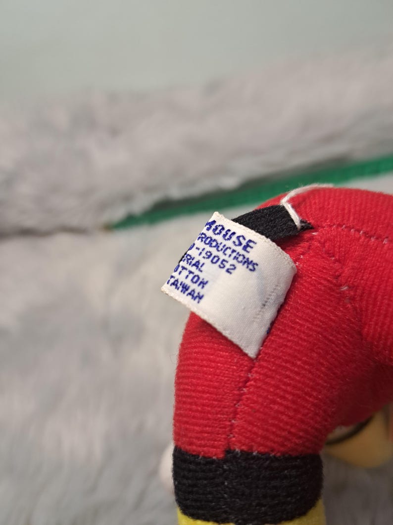Pu&ograve; includere: Primo piano del braccio rosso di un peluche con polsino nero e finiture gialle. Un'etichetta bianca &egrave; attaccata, che mostra il testo tra cui "HOUSE PRODUCTIONS" e "MATERIAL COTTON TAIWAN". Lo sfondo &egrave; un grigio morbido e sfocato.