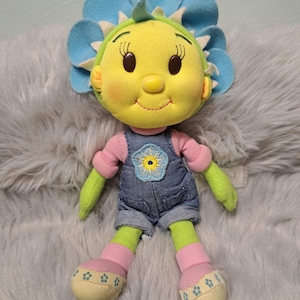 Puede incluir: Muñeca de peluche con cabeza en forma de flor y cara sonriente. La muñeca tiene cara amarilla, pétalos azules y brazos y piernas verdes. Viste un peto vaquero con un parche de flor y mangas y calcetines rosas. Los pies de la muñeca son de color crema con detalles florales.