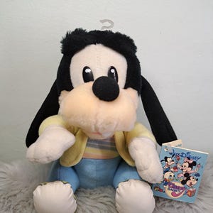May include: A plush Goofy character toy with black fur, a peach-coloured snout, and a single white tooth. The toy wears a yellow jacket over a pastel striped shirt and blue trousers. A Disney Babies tag is attached, featuring illustrations of Disney characters.