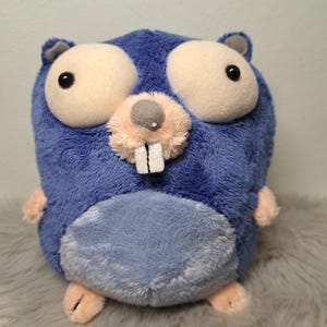 Puede incluir: Un peluche con forma de tuza con pelaje azul, grandes ojos blancos con pupilas negras, un hocico rosa claro y dos prominentes dientes delanteros blancos. La criatura tiene pequeñas patas de color rosa claro y un cuerpo redondeado con una mancha azul claro.