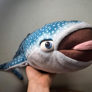 May include: A plush whale shark toy with a blue body covered in white spots. The toy has a large, open mouth with a brown interior and a pink tongue. The whale shark has a large, expressive eye.