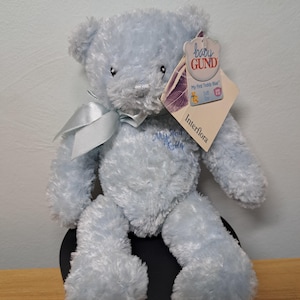 May include: A pale blue teddy bear with a satin bow. The bear has a tag that reads "baby GUND My First Teddy Blue". The bear is sitting on a black surface.