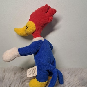 May include: A plush Woody Woodpecker toy featuring a bright red crest, a yellow beak with black stitched details, a blue body, white gloves, and yellow feet. A small white fabric tag with printed text is attached to the side of the blue torso.