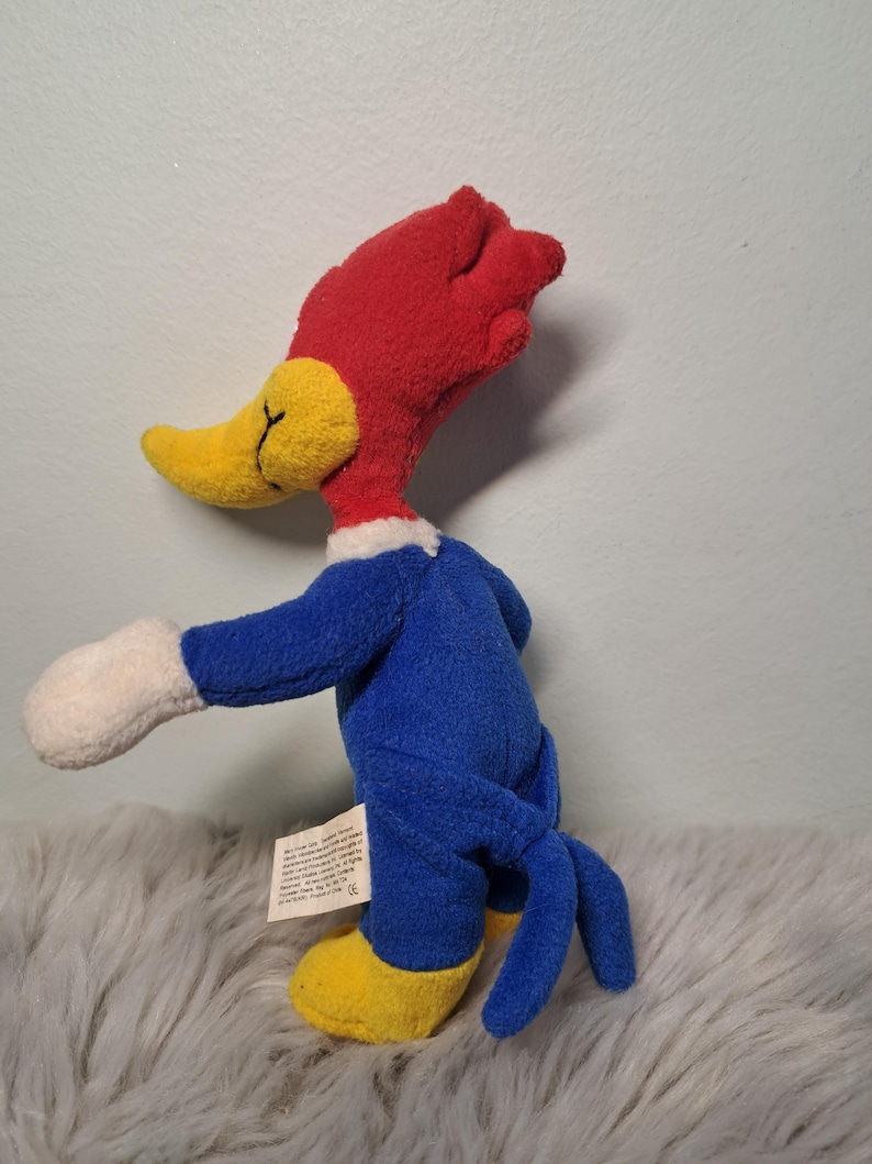 May include: A plush Woody Woodpecker toy featuring a bright red crest, a yellow beak with black stitched details, a blue body, white gloves, and yellow feet. A small white fabric tag with printed text is attached to the side of the blue torso.
