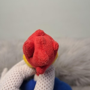 May include: A plush toy featuring a bright red textured head with a yellow accent at the base, attached to a blue body. The material appears soft and fuzzy, with a distinct rounded shape at the top of the head and a blue limb visible below.