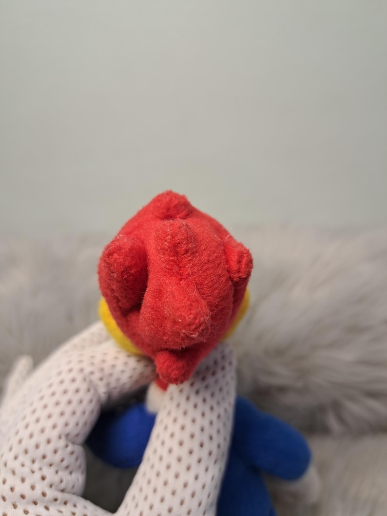 May include: A plush toy featuring a bright red textured head with a yellow accent at the base, attached to a blue body. The material appears soft and fuzzy, with a distinct rounded shape at the top of the head and a blue limb visible below.