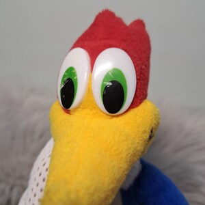 May include: A plush toy character featuring a bright red crest, a prominent yellow beak, and large white oval eyes with green irises and black pupils. The character has blue fabric on the sides and white mesh-like fabric on the lower body section.
