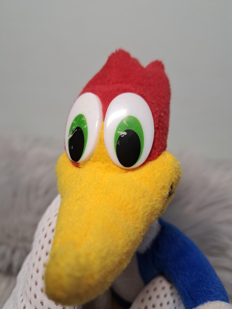 May include: A plush toy character featuring a bright red crest, a prominent yellow beak, and large white oval eyes with green irises and black pupils. The character has blue fabric on the sides and white mesh-like fabric on the lower body section.